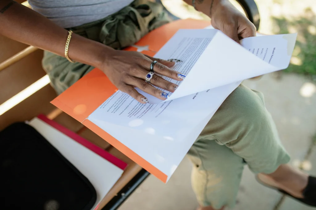 A person sitting on a bench flips through printed documents in an orange folder, wearing green pants and jewelry on their hands.